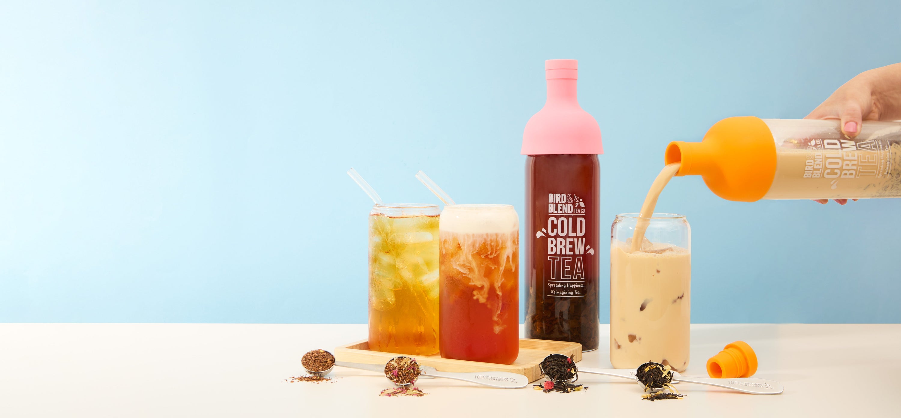 cold brew tea bottles peachy pink filled with tea & cups of cold brew iced tea & spoons filled with loose leaf tea showcasing cold brew teas in lemonade water & milk 