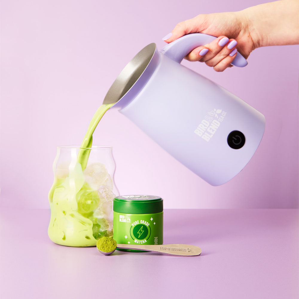 soft lilac magic whisk pouring pure grade matcha tea into ice in a cup with matcha tea tin and spoon